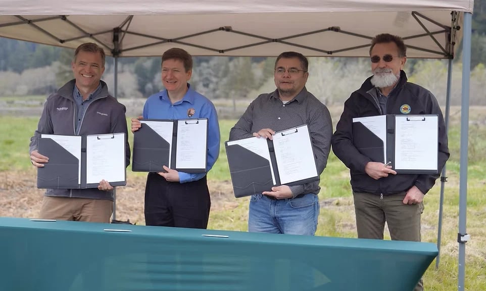 Four men in casual attire holding certificates under a tent at an outdoor event with green field in background