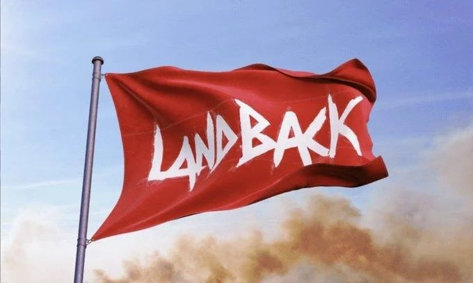 Red flag with white LAND BACK text waving on a pole against blue sky with dust clouds