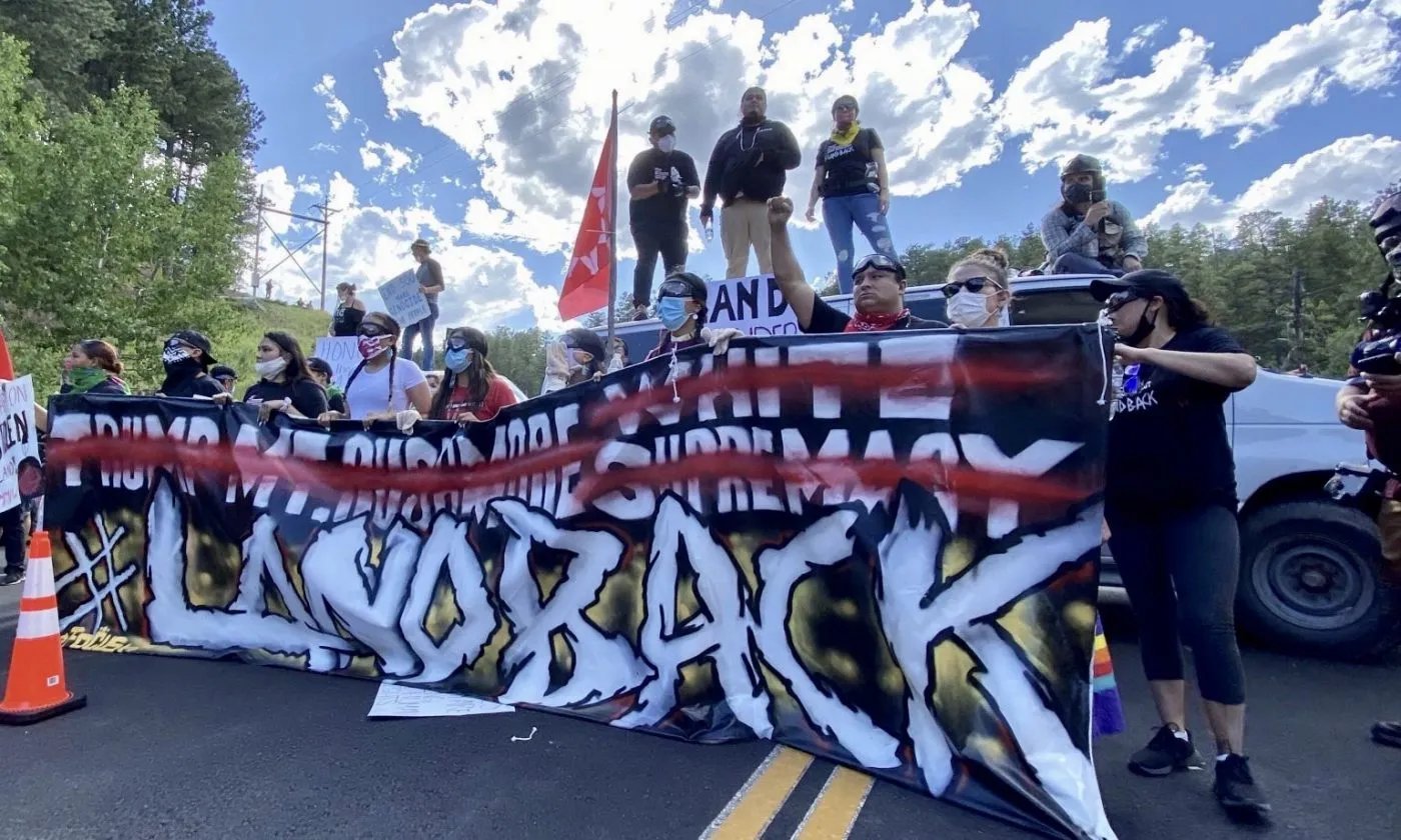 Group of people at outdoor rally holding large banner with graffiti-style text on pickup truck under blue sky