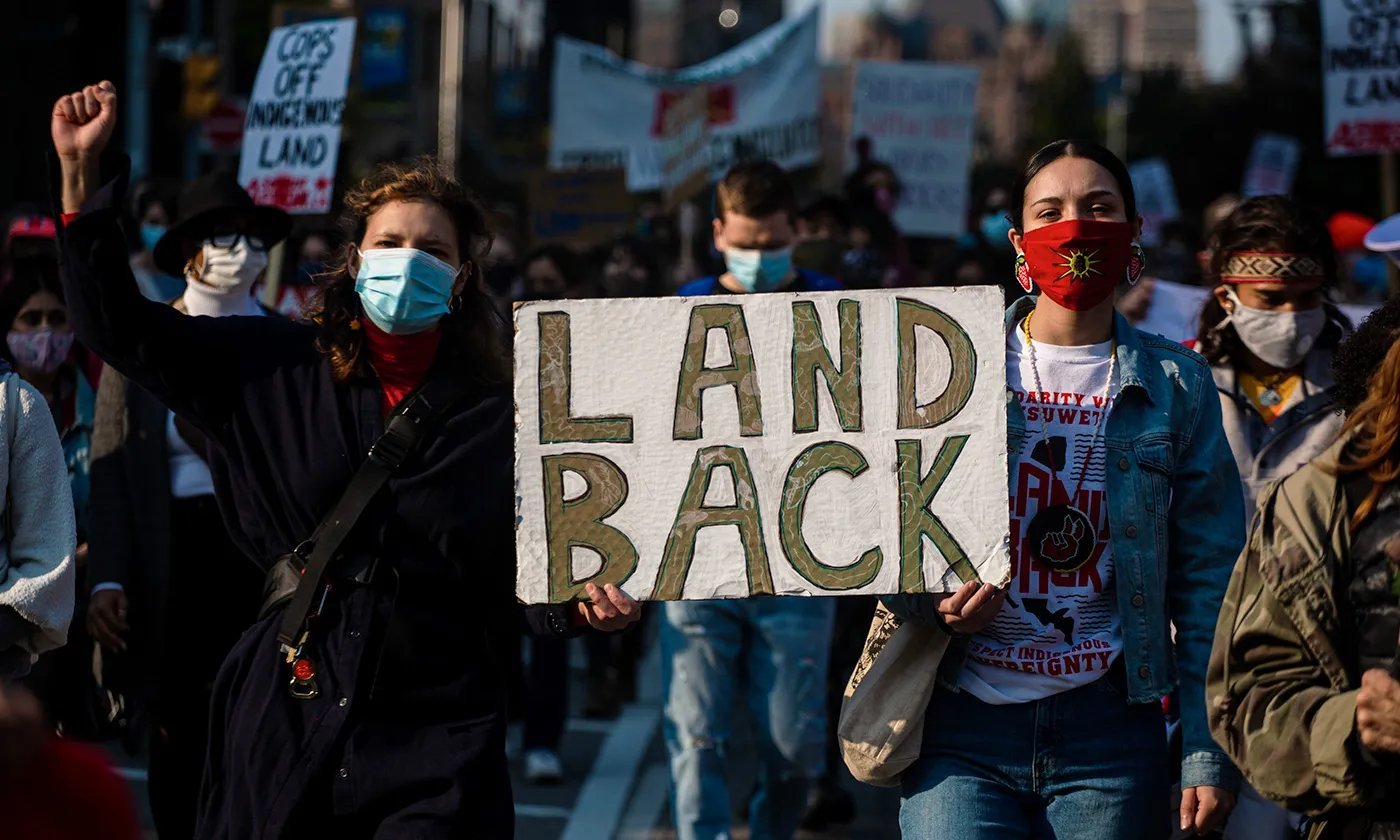 Protesters in masks hold a LAND BACK sign at a demonstration with other picketers visible in the background