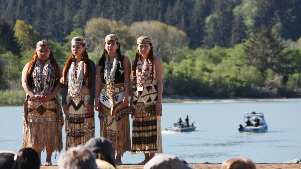 Four Indigenous women in traditional regalia stand on a rocky beach with forest and water in the background